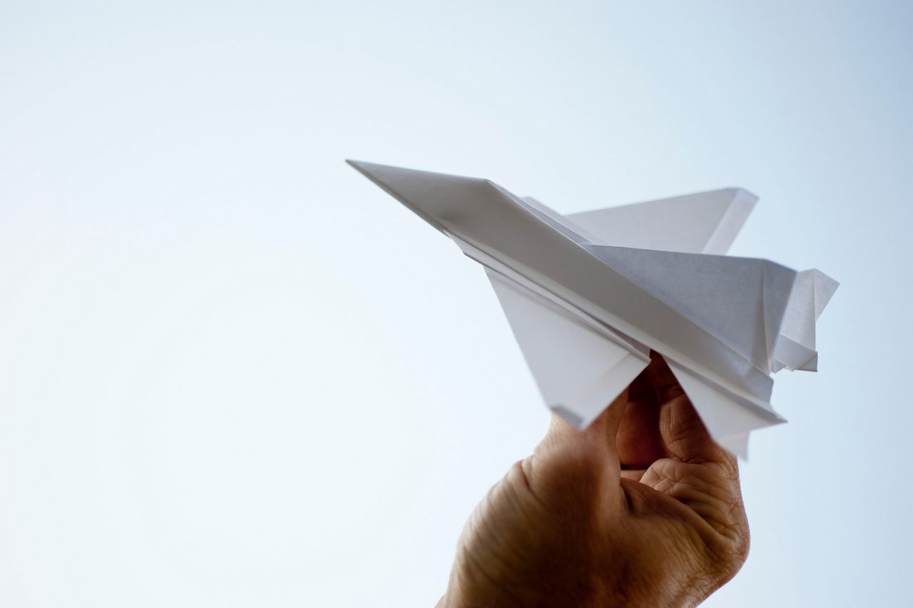 A hand holds a folded paper airplane against a clear sky, symbolizing accessible air travel planning.
