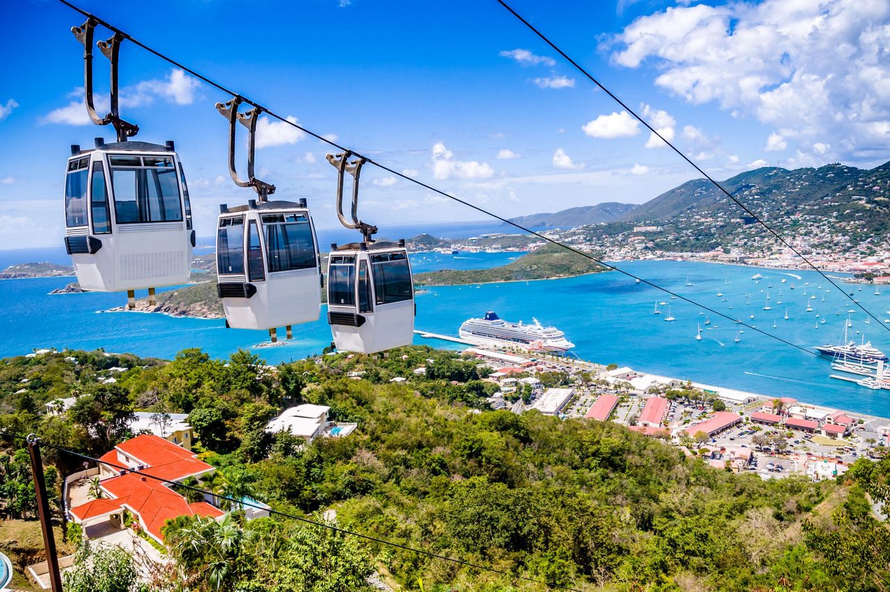 White cable car gondolas suspended over tropical landscape with cruise ship in harbor and mountainous coastline in background
