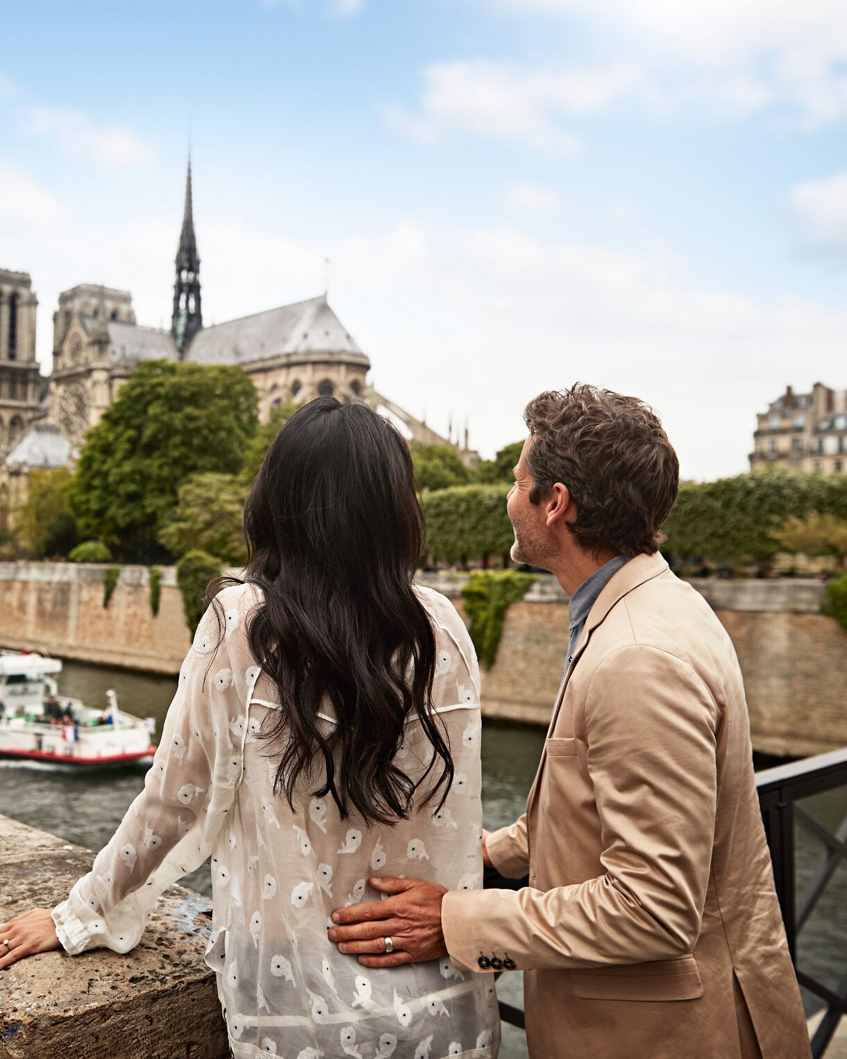 A couple stands on a bridge overlooking the Seine River in Paris, gazing at the iconic Notre-Dame Cathedral. The man gently places his hand on the woman's back as they enjoy the scenic view, with trees and boats visible along the river.