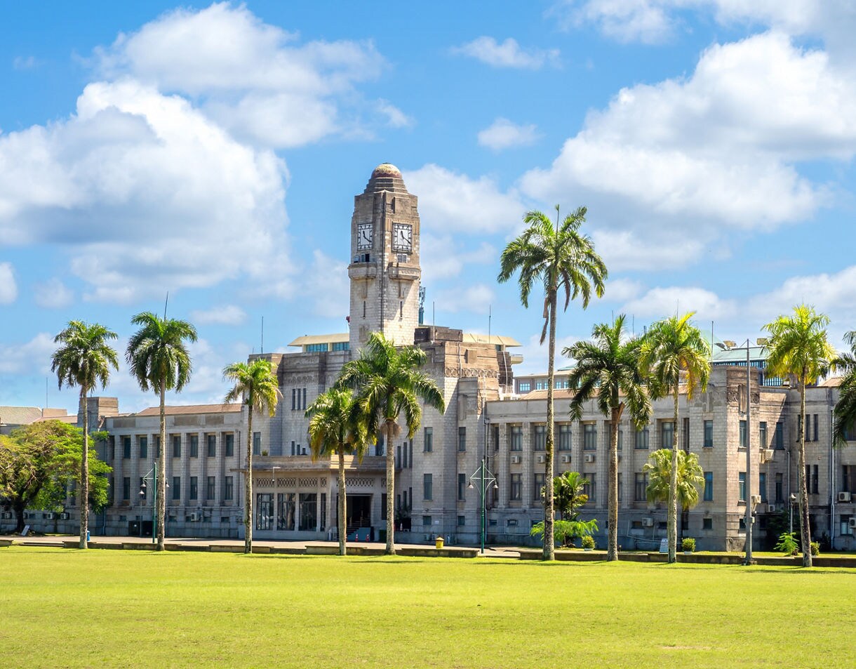 Historic stone parliament building with a central clock tower, lined with tall palm trees and set against a bright blue sky.