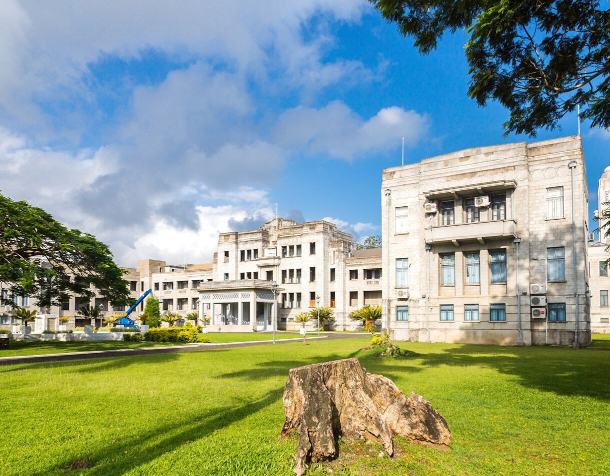 Wide view of Fiji’s Parliament House, a pale stone building with columns and tall windows, surrounded by green grass and trees under a partly cloudy blue sky.