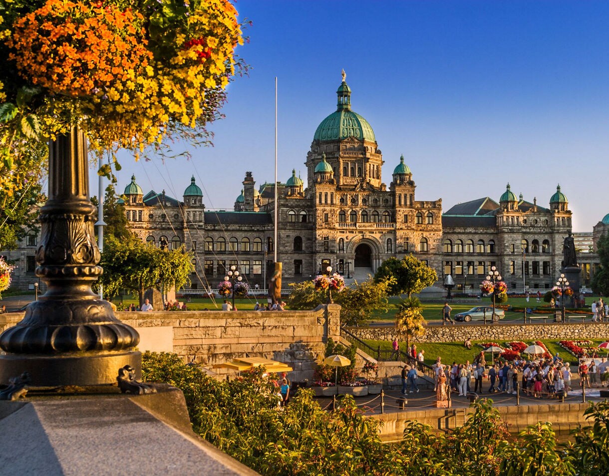 British Columbia Parliament Buildings with distinctive green copper domes bathed in warm golden light during sunset, with people walking on the grounds and ornate lamp posts in the foreground