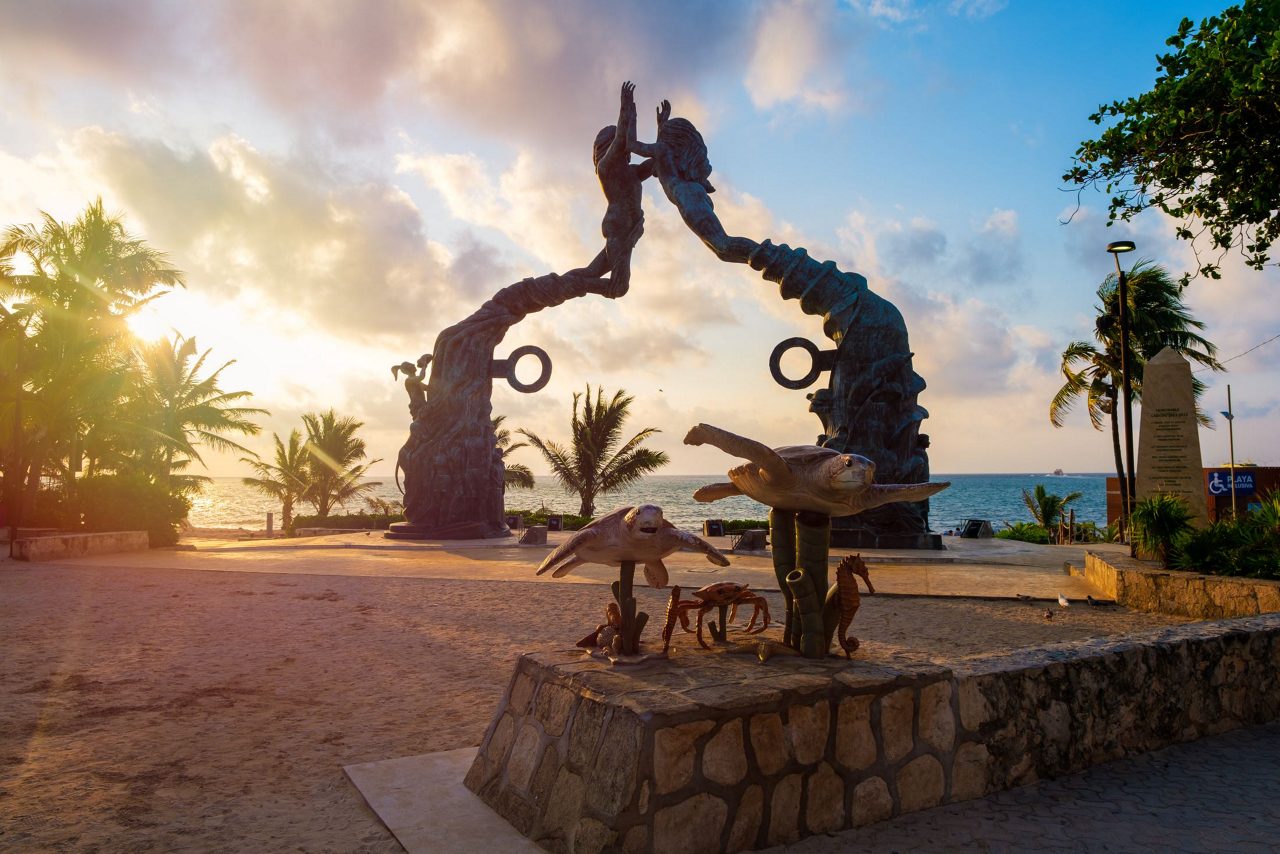 Portal Maya monument featuring sculptured figures forming an arch at sunset on Cozumel beach with palm trees and Caribbean Sea in background.