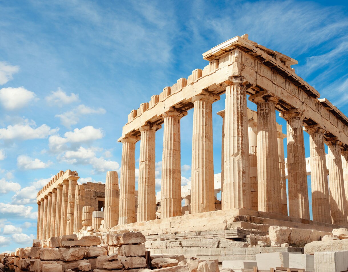 The Parthenon on the Acropolis in Athens, showing tall stone columns and weathered ruins beneath a bright blue sky with scattered clouds.