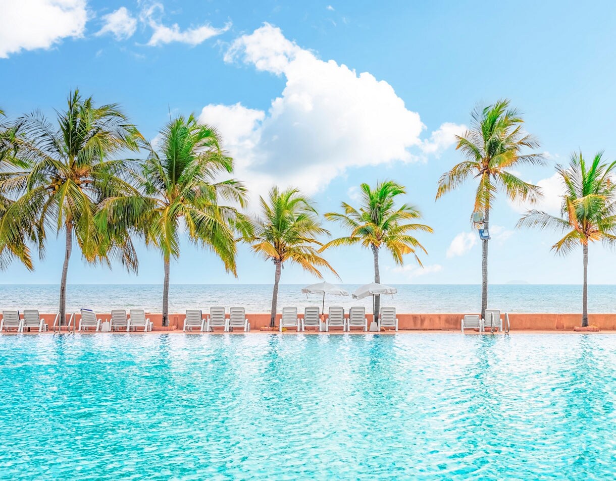 Sparkling blue swimming pool lined with lounge chairs and umbrellas, with tall palm trees and the sea in the background.