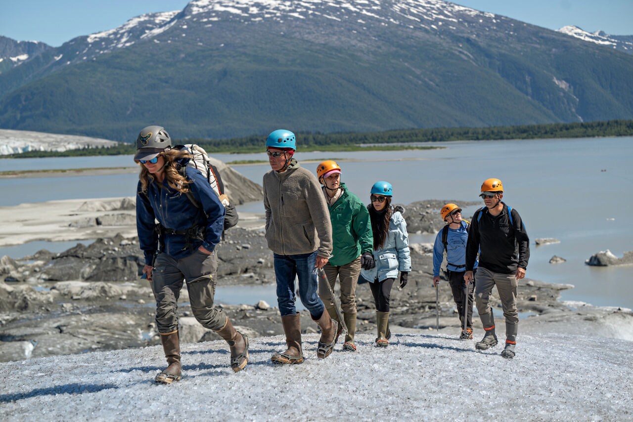 A group of adult hikers walks in a line across a glacier or snowy terrain.
