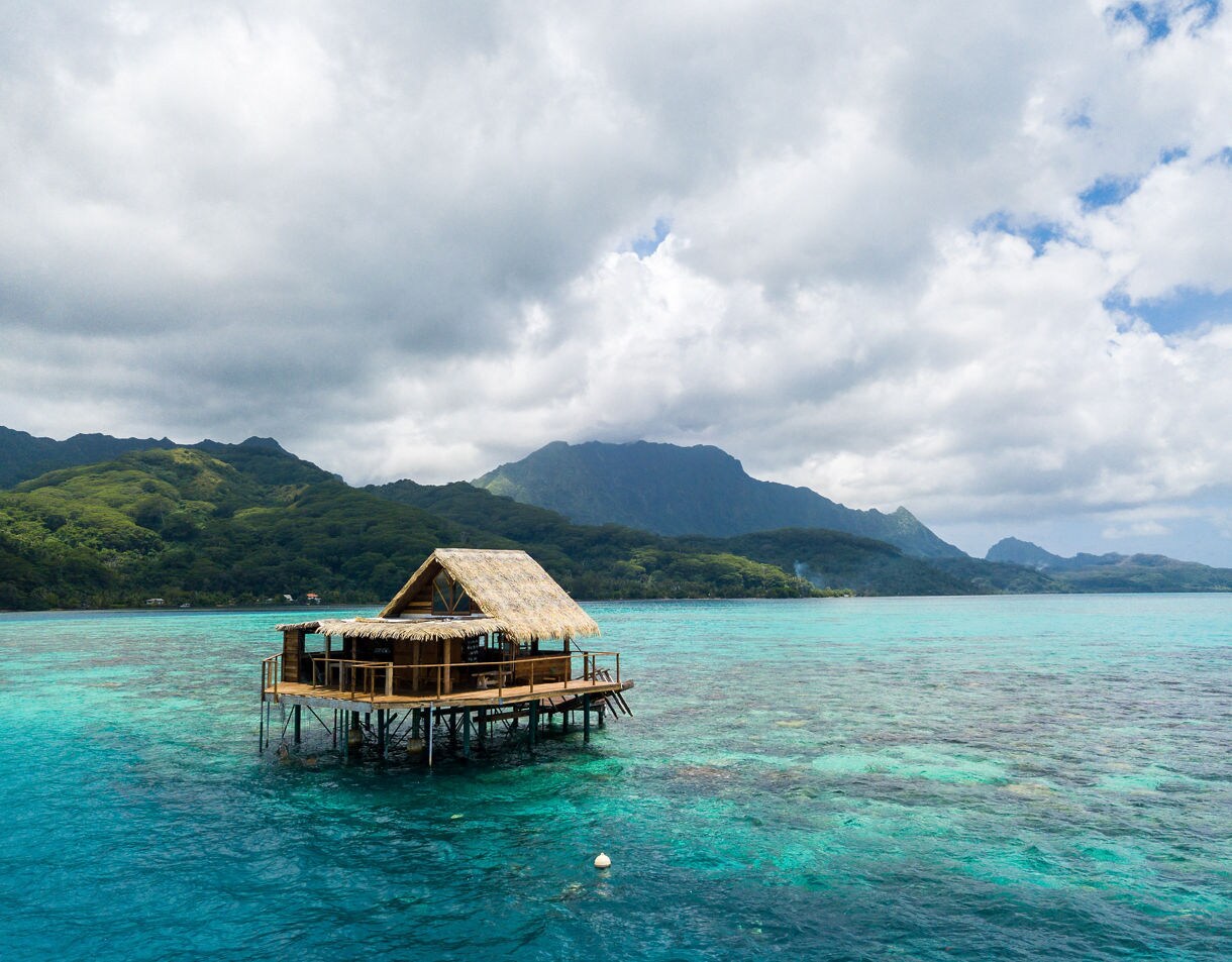 Wooden thatched-roof hut on stilts over clear turquoise water, with Raiatea’s green mountains and cloudy sky in the background.