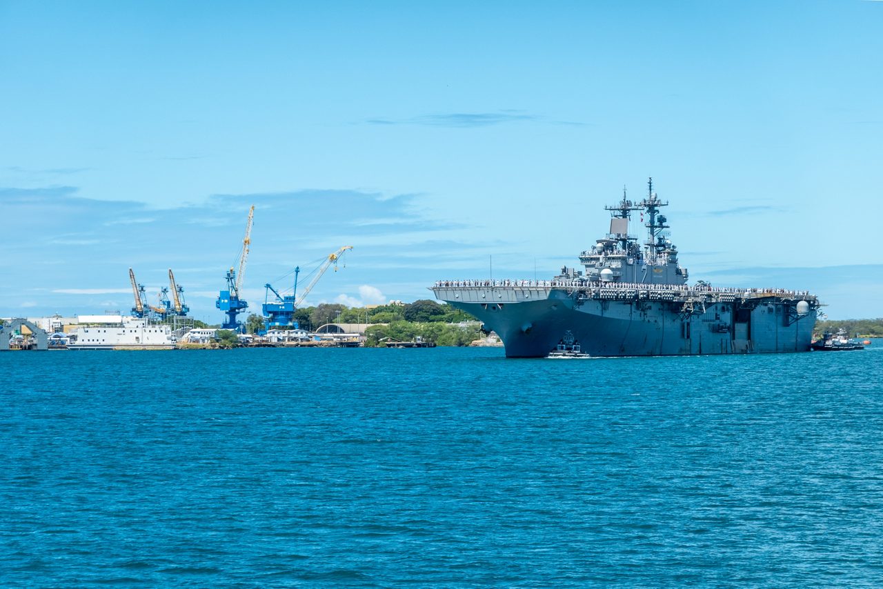 A large U.S. Navy ship docked in Pearl Harbor with cranes and facilities in the background, under clear blue skies.