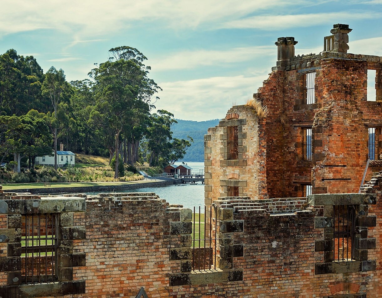 Weathered brick walls of the Port Arthur penitentiary overlooking calm water and a forested shoreline in Tasmania.
