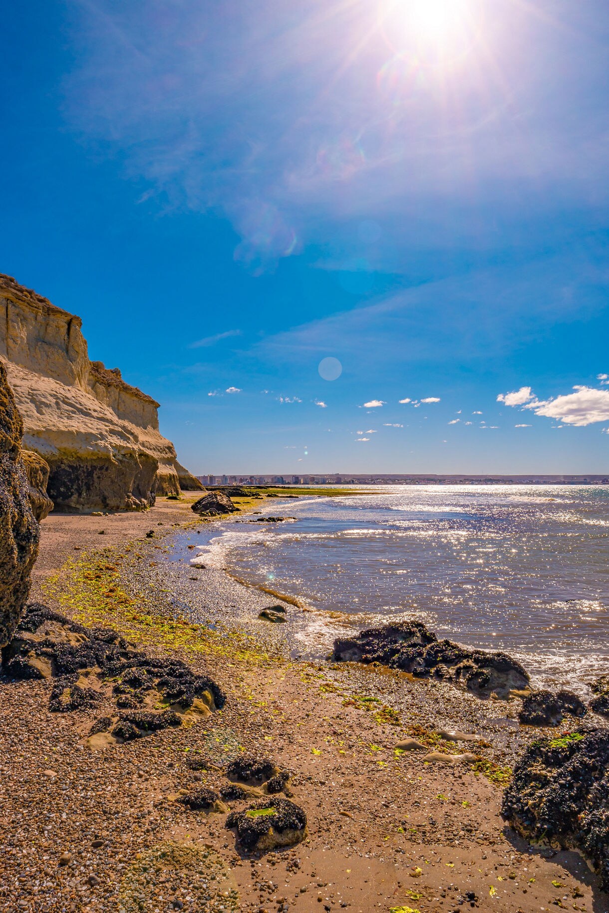 A bright coastal scene with golden cliffs, a rocky shoreline and sparkling blue water under strong midday sun.
