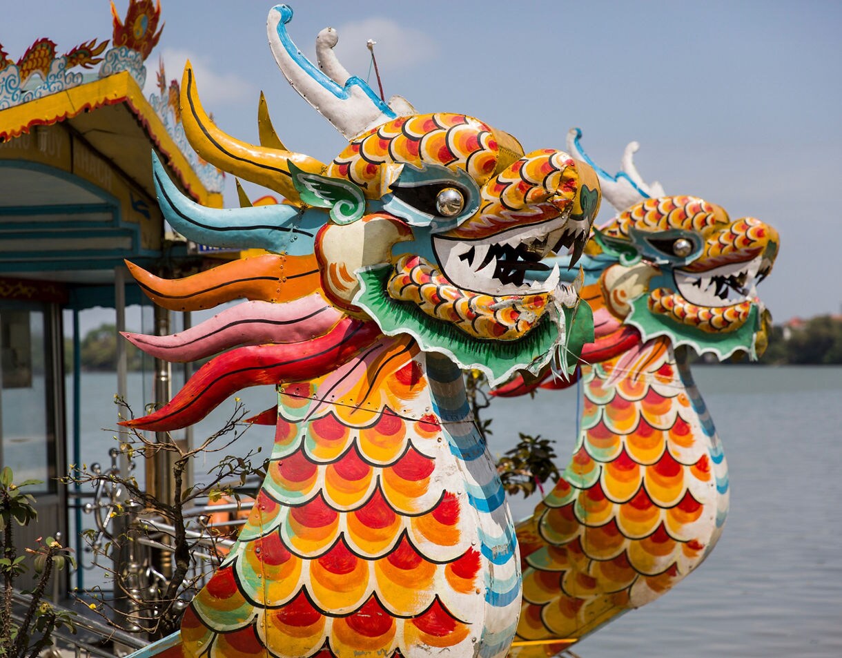 Close-up of two brightly painted dragon boat heads on the Perfume River in Hue, Vietnam, featuring vivid scales, colorful manes and ornate detailing under the sun.