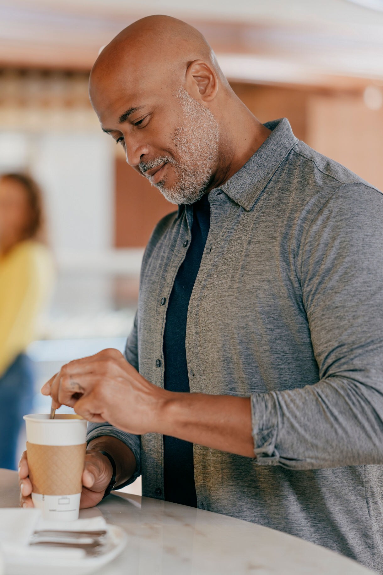 Person adding cream to coffee at casual café counter with pastry display case in background.