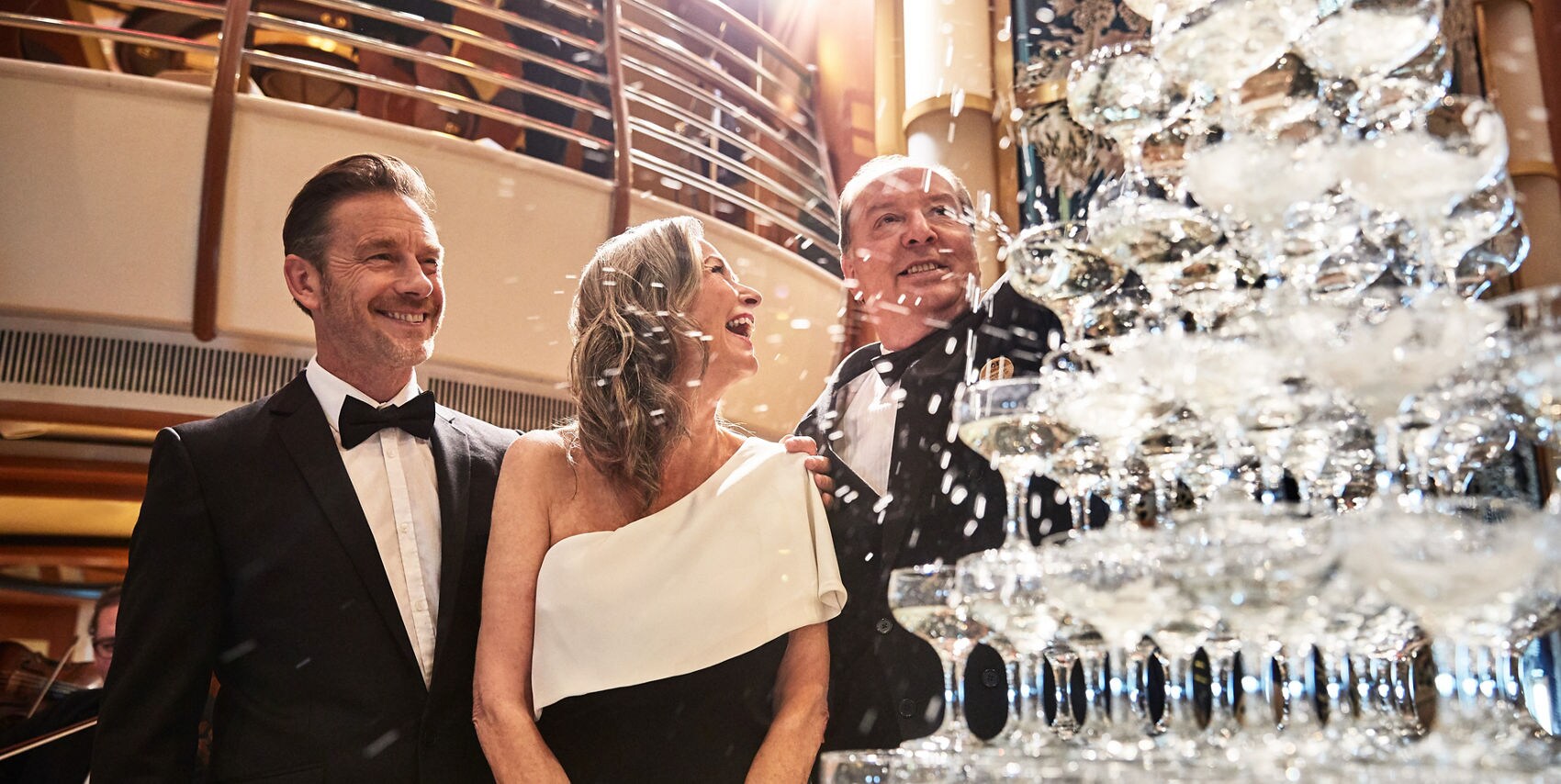 Three adults dressed in formal evening attire stand beside a tall champagne glass tower. The setting appears to be a luxurious indoor venue with warm lighting and stylish architecture. The mood is celebratory and sophisticated, with sparkling reflections from the glass pyramid.