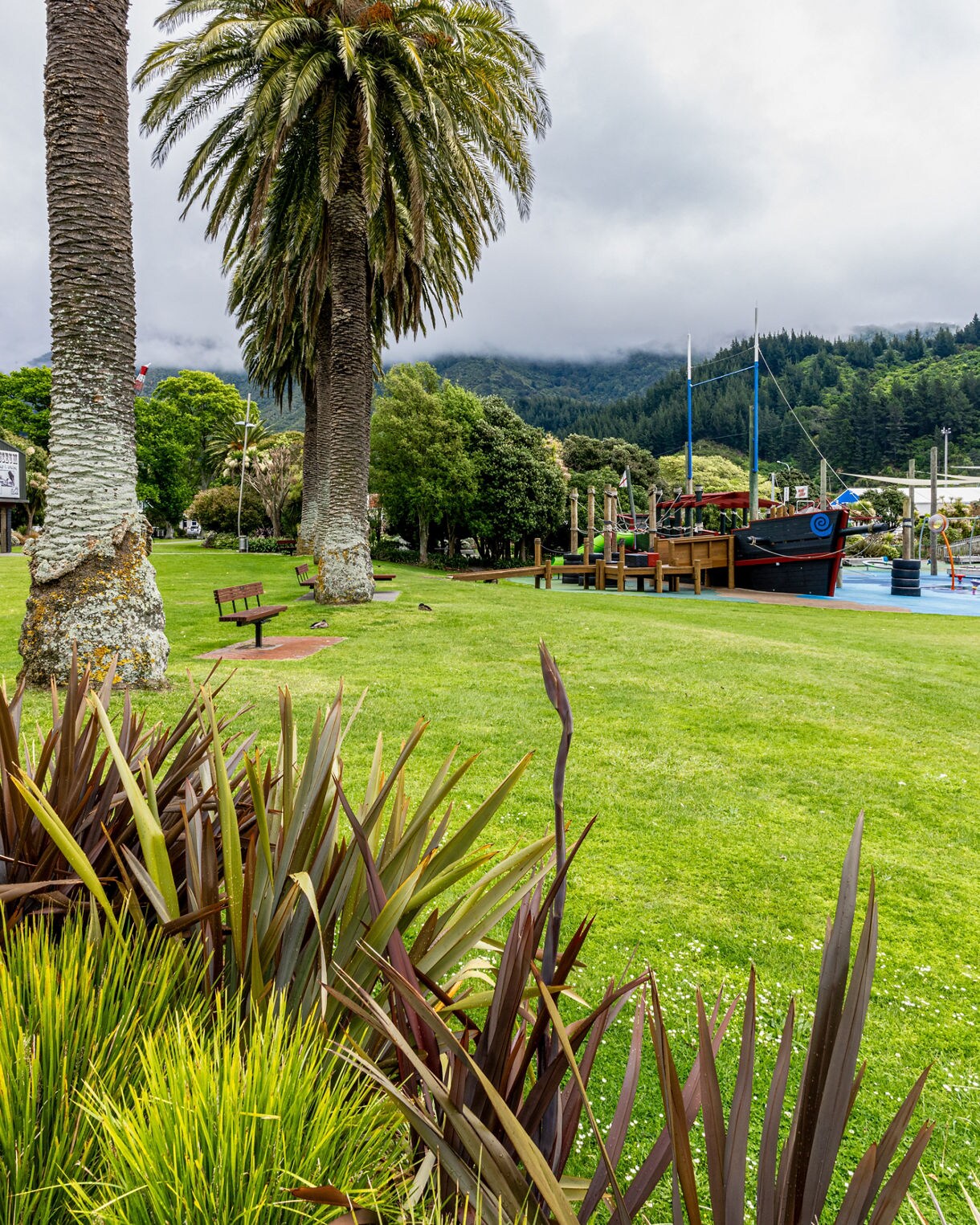 Green park area in Picton with tall palm trees, trimmed grass and a colorful playground ship, with forested hills rising misty in the background..