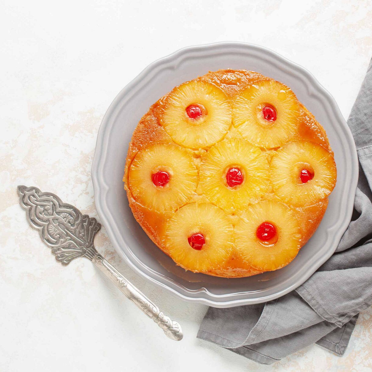 Overhead view of a pineapple upside-down cake topped with caramelized pineapple rings and maraschino cherries, served on a gray plate with a vintage cake server and gray napkin.