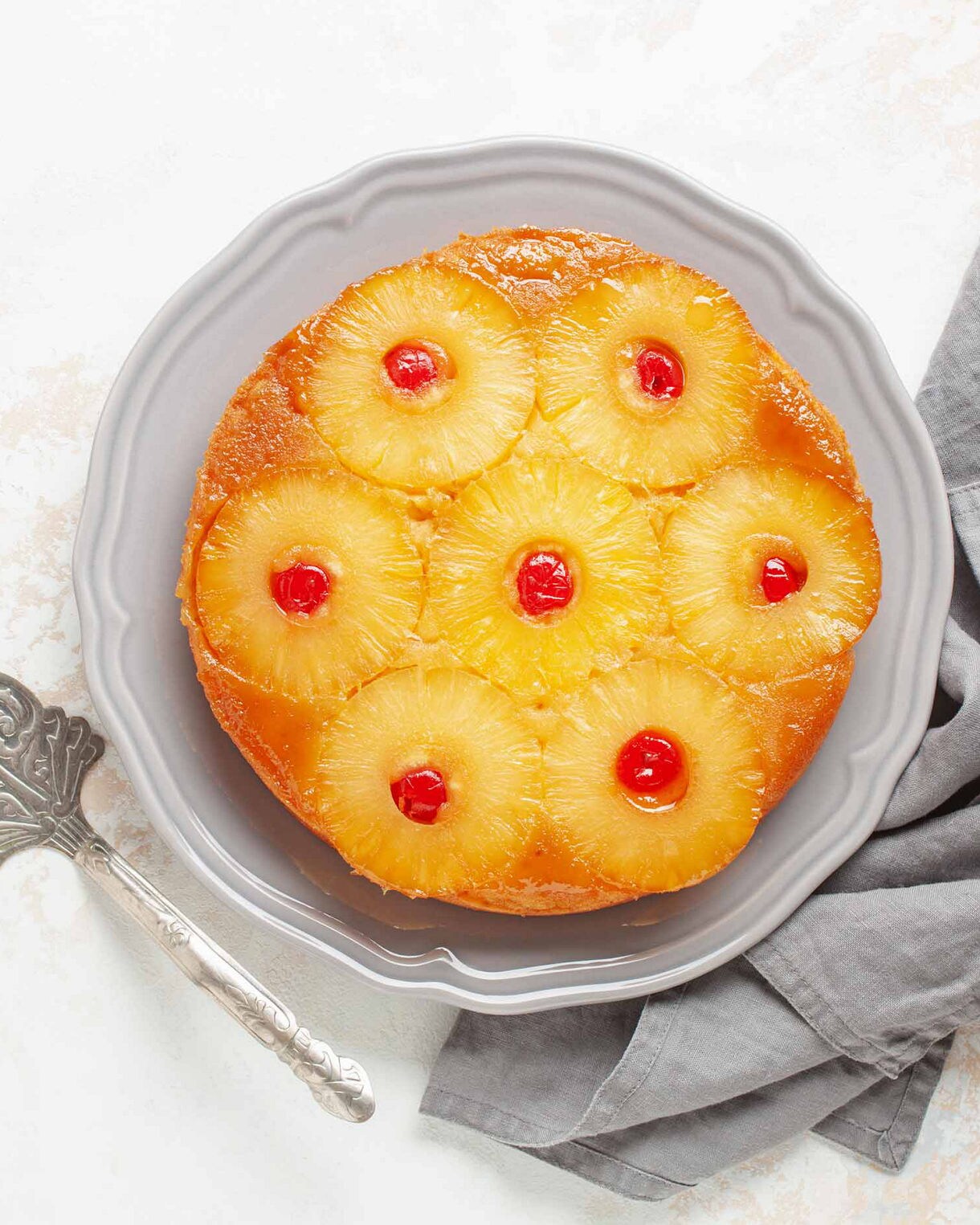 Overhead view of a pineapple upside-down cake topped with caramelized pineapple rings and maraschino cherries, served on a gray plate with a vintage cake server and gray napkin.