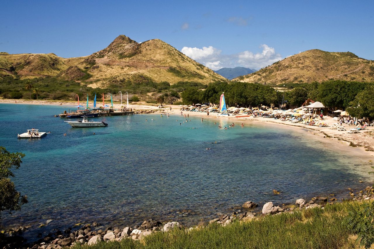 Aerial view of a tropical beach with clear blue water, white sand, beach umbrellas, boats, and volcanic hills in the background on St. Kitts and Nevis.