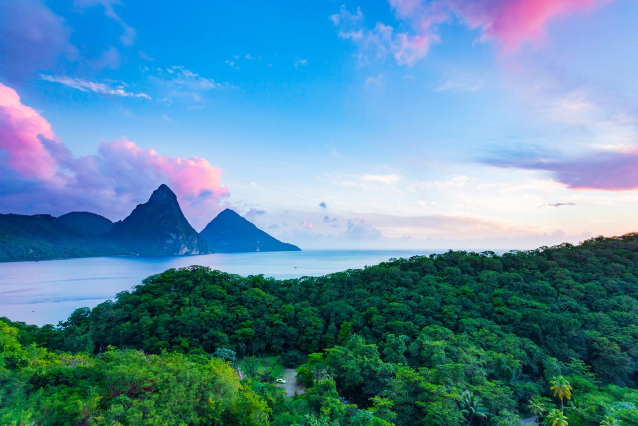 Aerial view of St. Lucia's famous twin Piton peaks jutting from the ocean with lush green rainforest in foreground under dramatic pink and blue sunset sky.