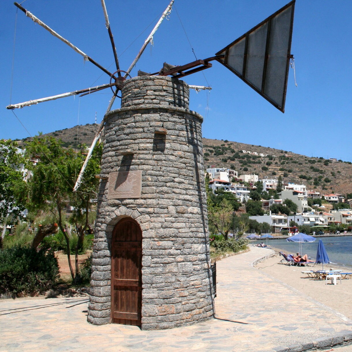 A stone windmill stands near a sandy beach in Elounda with lounge chairs and umbrellas set along the calm blue water and hillside homes in the distance.