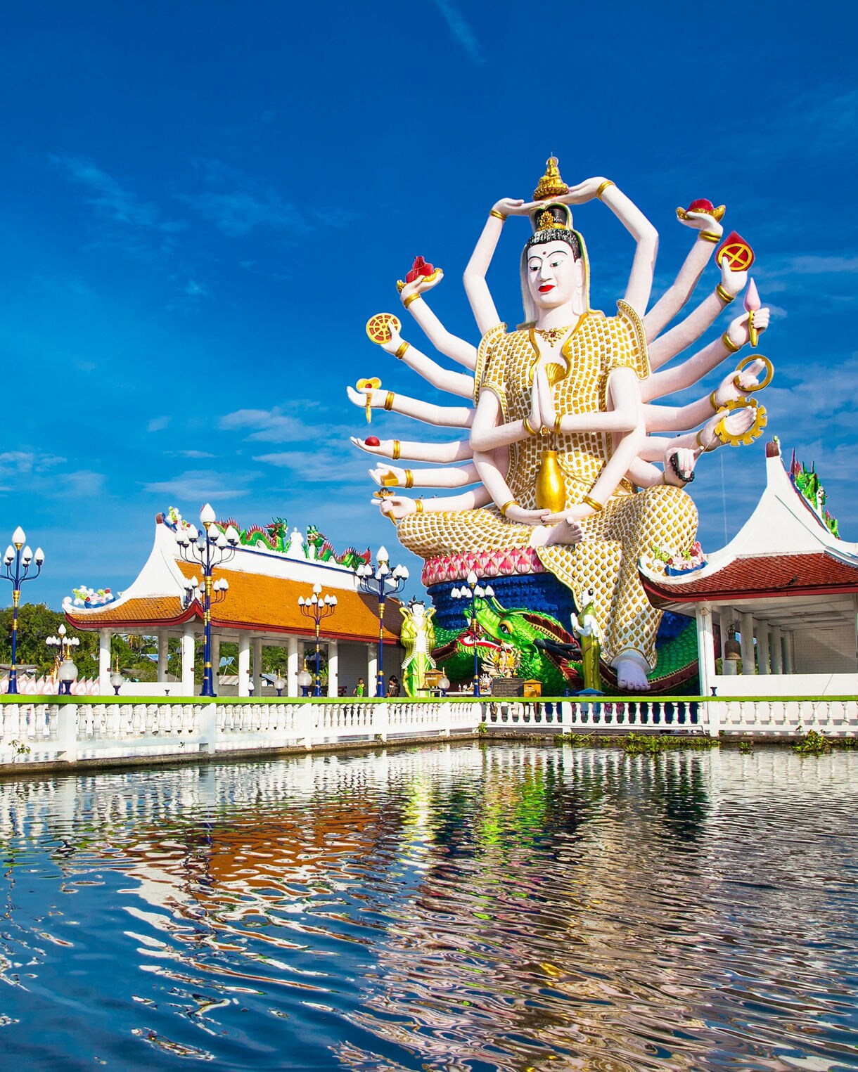 Plai Laem Temple in Koh Samui featuring a colorful 18-armed Guanyin statue beside ornate temple buildings reflected in a tranquil pond.