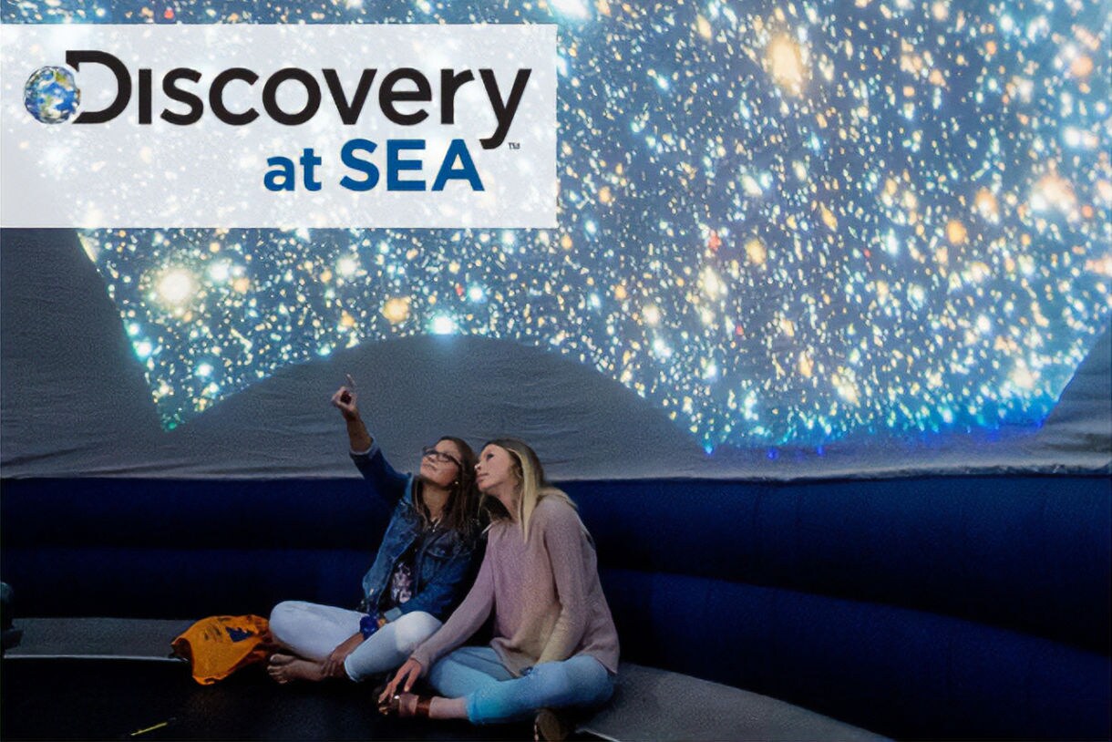 Two friends marvels at the night sky from the top deck of a Princess Cruises ship.  The indoor planetarium and the engaging Discovery™ Voyage program logo are featured in the background. [Courtesy Princess Cruises]