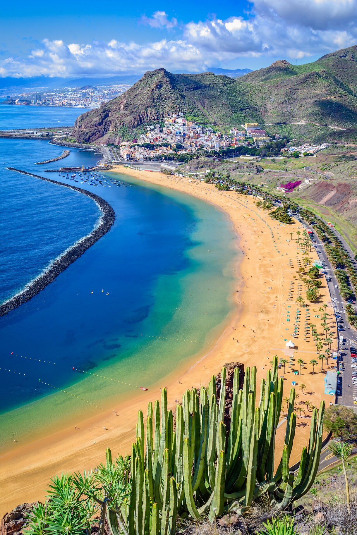 Aerial view of Playa de Las Teresitas in Tenerife with golden sand, turquoise water and lush green mountains rising behind the beach.