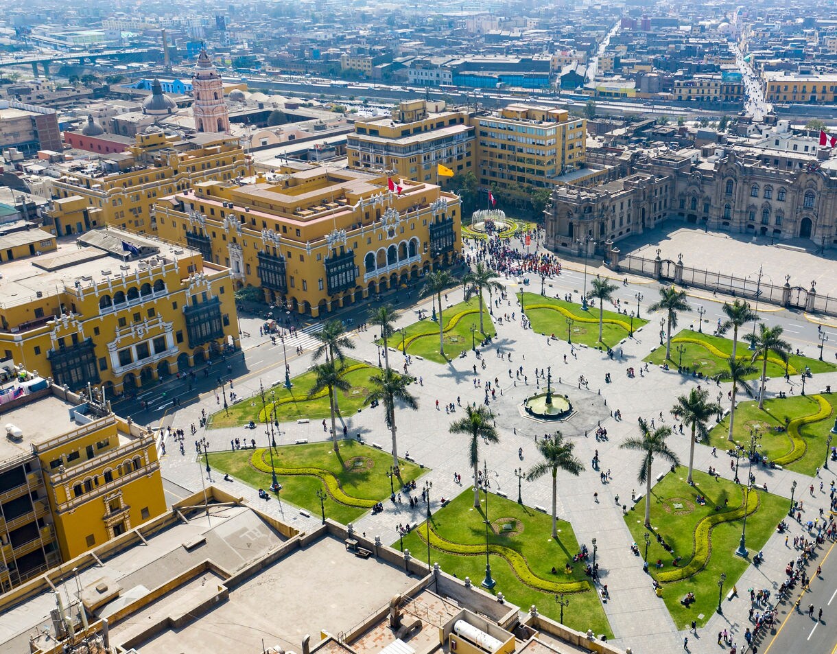 Aerial view of Plaza de Armas in Lima showing a large square with palm-lined walkways, a central fountain, yellow colonial buildings and the Presidential Palace surrounded by busy streets.