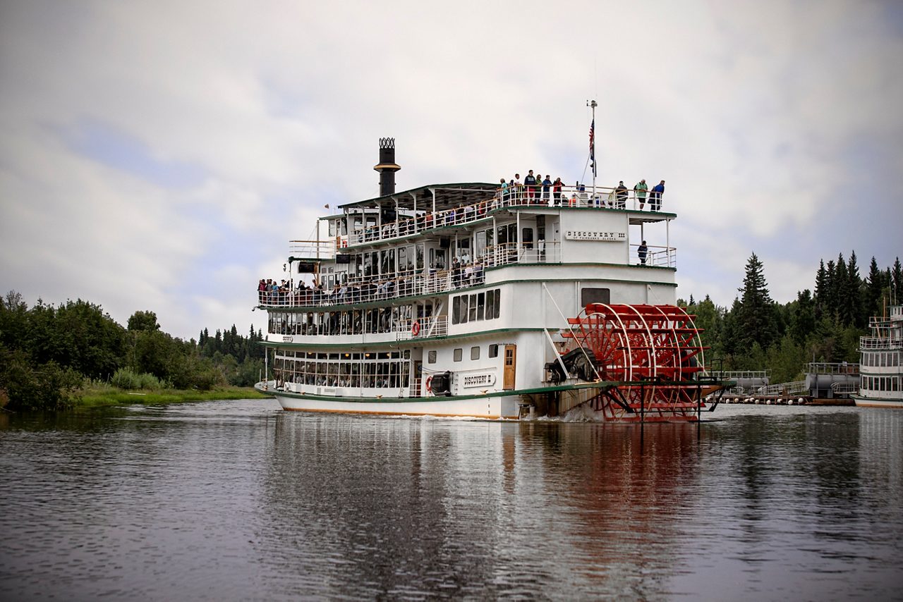 Restored Canadian Pacific sternwheeler docked beside a lake, with a red paddlewheel, wooden walkway, picnic tables, and forested mountains in the background