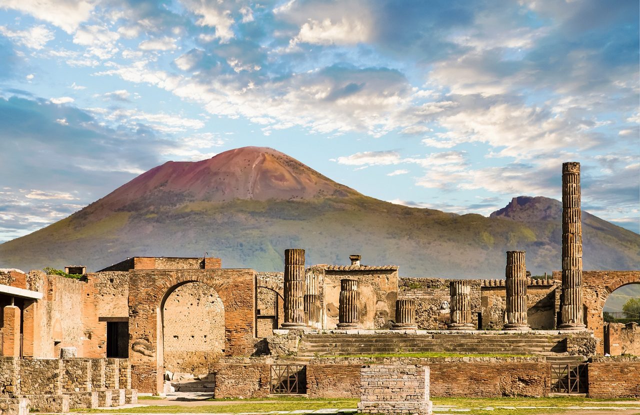 Ruins of Pompeii with crumbling stone columns and brick walls, set against the backdrop of Mount Vesuvius under a partly cloudy sky.