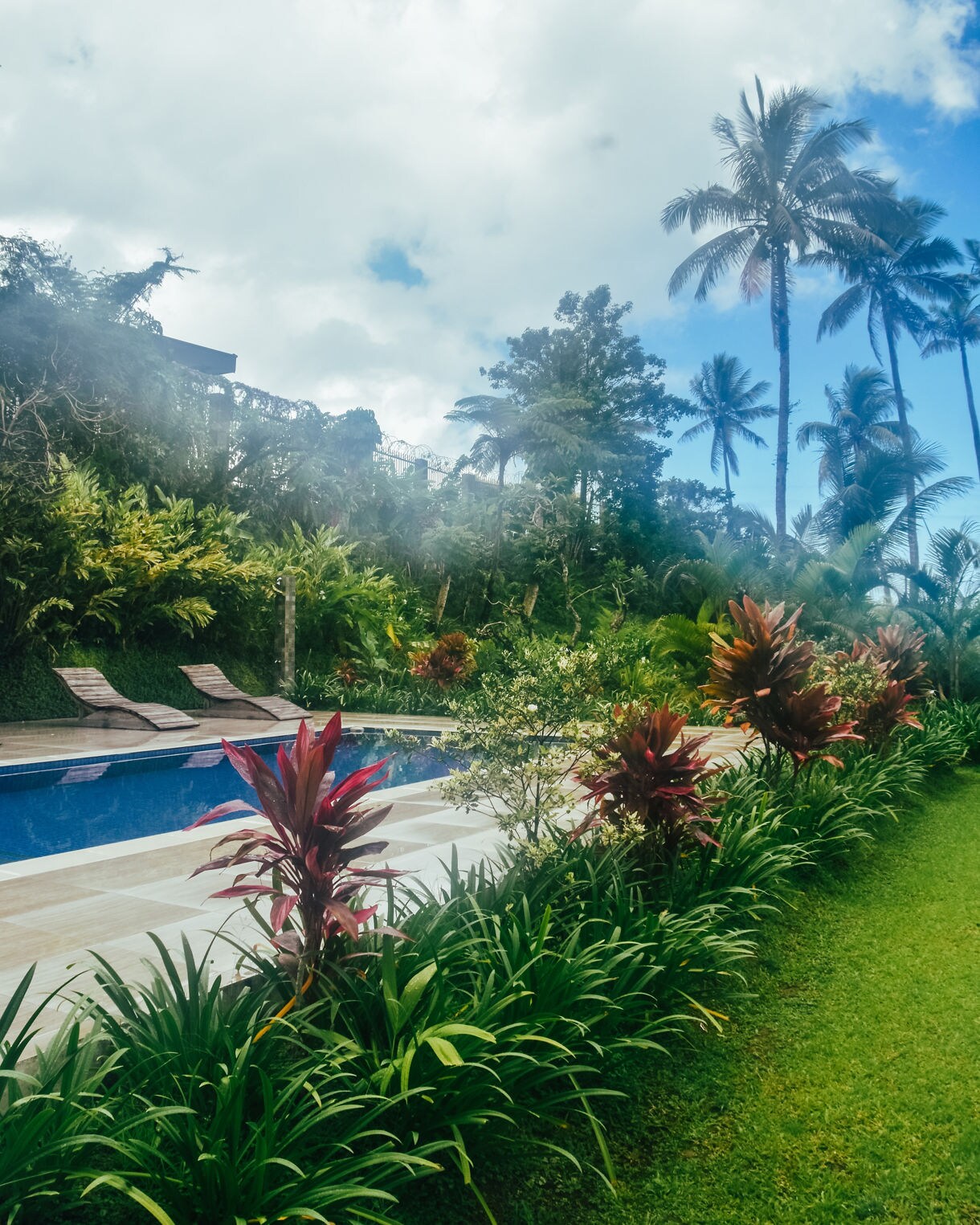 A serene outdoor pool bordered by lush tropical plants with tall palm trees and a cloudy blue sky overhead.