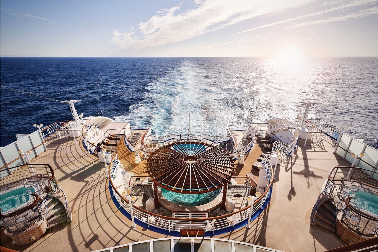 A stunning view from the deck of a Princess Cruises ship, showcasing a luxurious outdoor area with hot tubs, loungers, and a central shaded seating area. The ship's wake trails behind in the open ocean under a bright, clear sky, with the sun low on the horizon. 