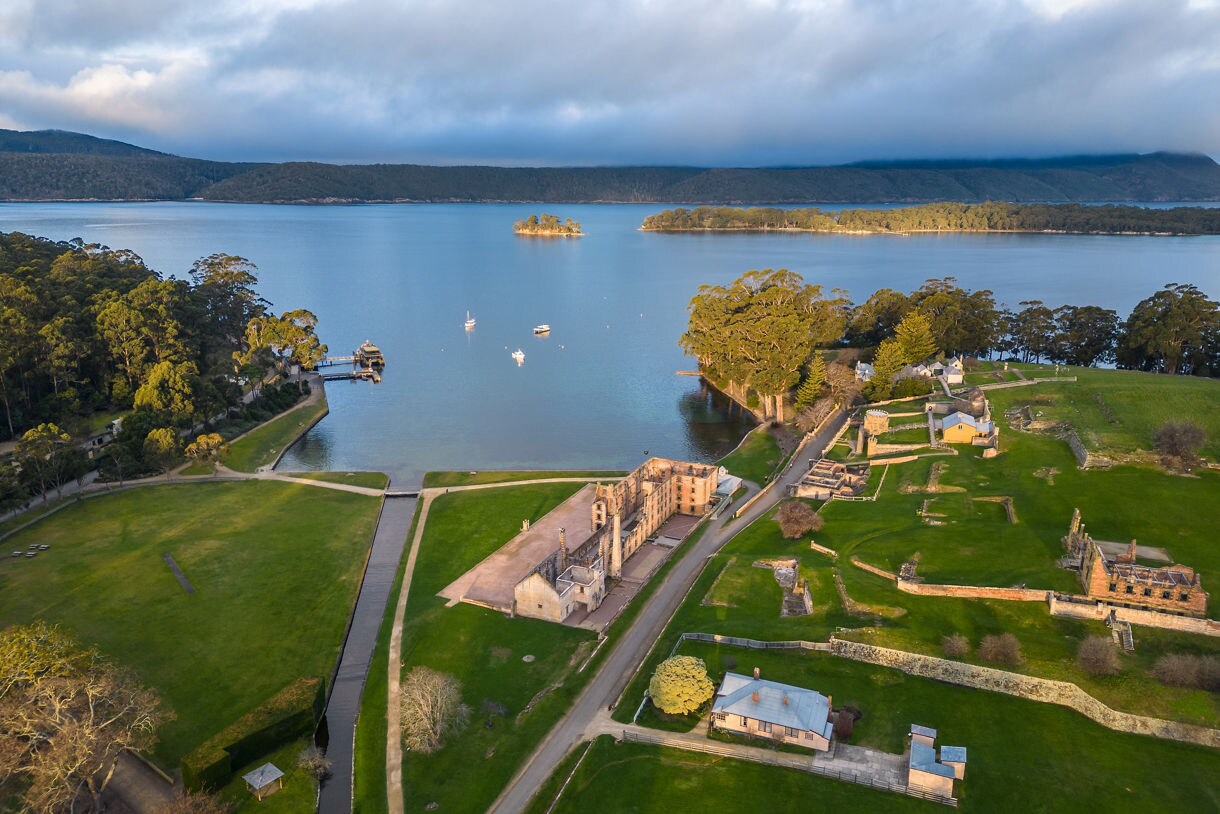 Aerial view of Port Arthur’s historic ruins beside a quiet blue harbor, surrounded by grassy grounds, forests and distant hills under soft morning light.