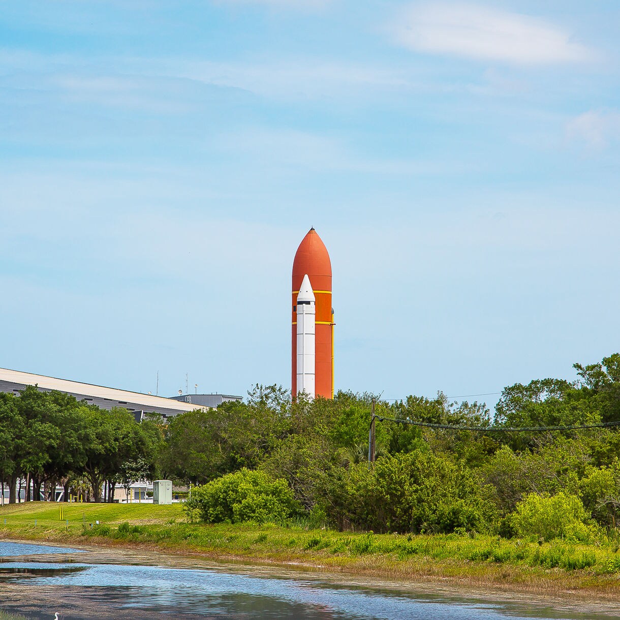 A space shuttle replica with an orange external tank is displayed outdoors near a lush green landscape and a waterway. The setting includes a modern building in the background under a bright blue sky. The scene captures a mix of technology and nature in a serene environment.