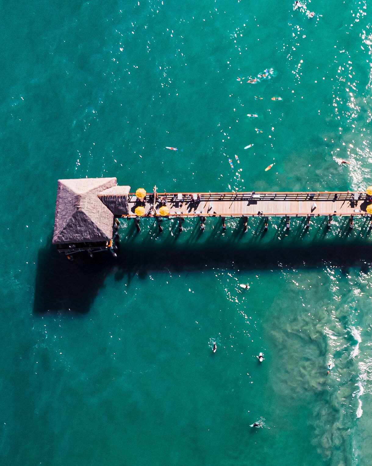 Aerial view of a pier and beach scene with a thatched roof building, yellow umbrellas, and people enjoying the ocean. 