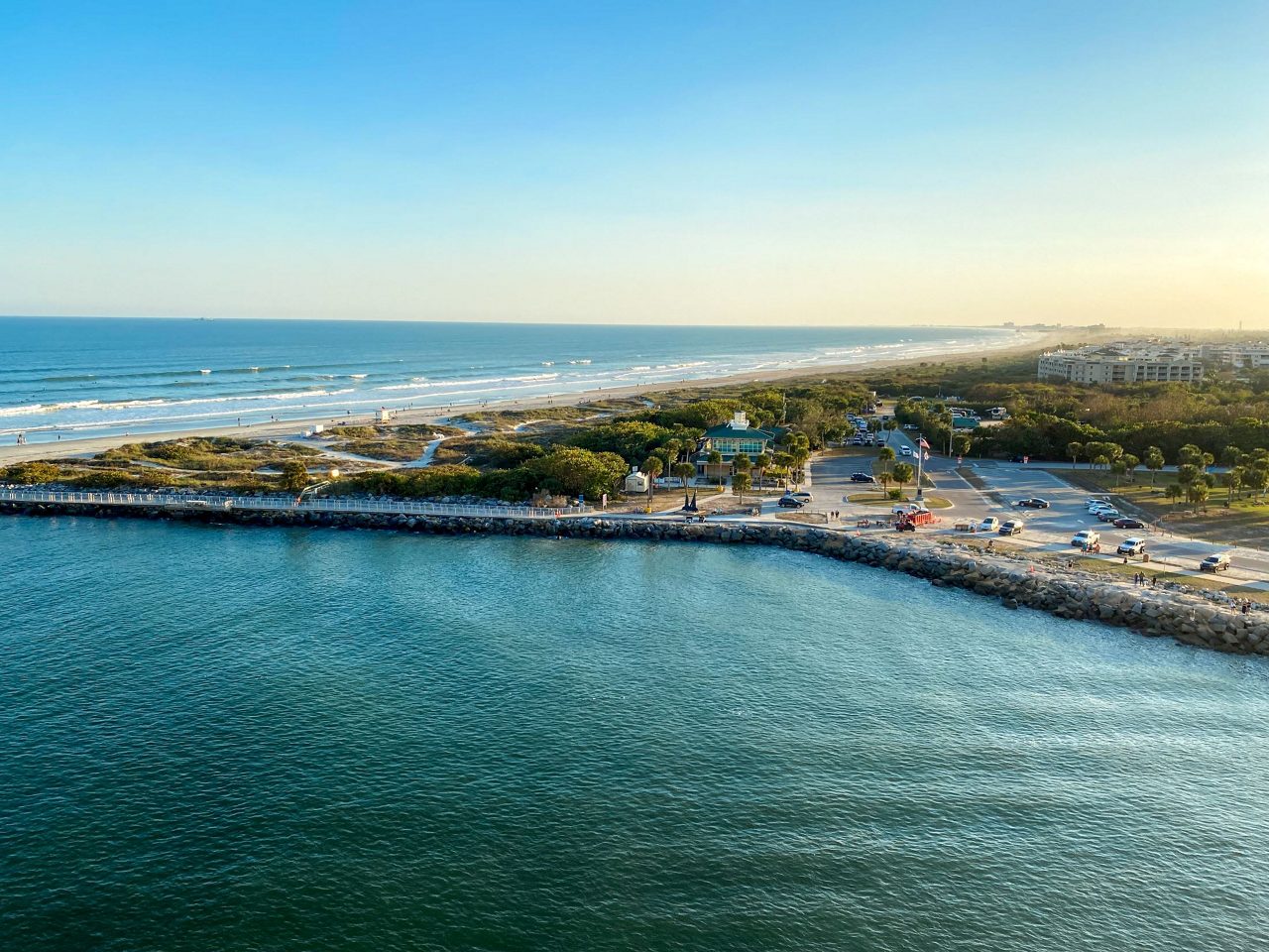 Aerial view of a beach with a parking lot and road along the coastline, surrounded by trees and bushes.