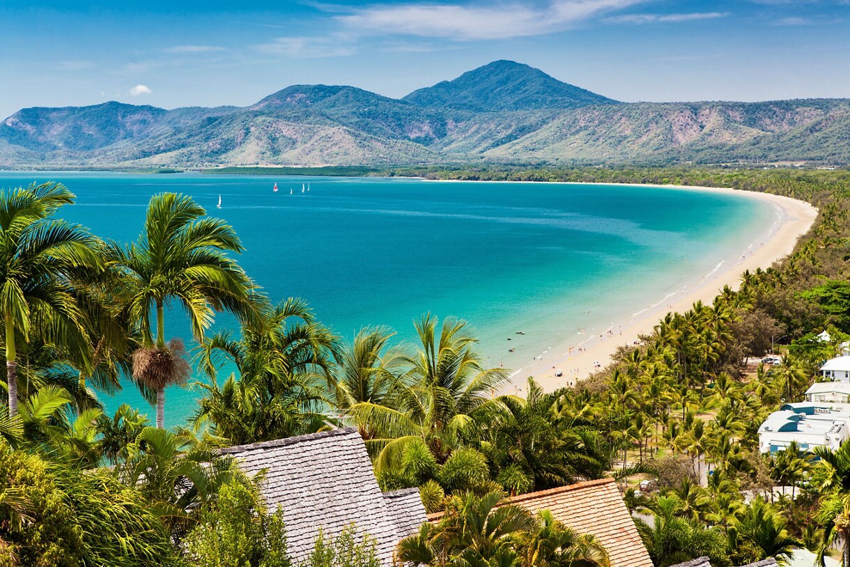 A long curved beach with turquoise water, lush palm trees and green mountains in the distance.