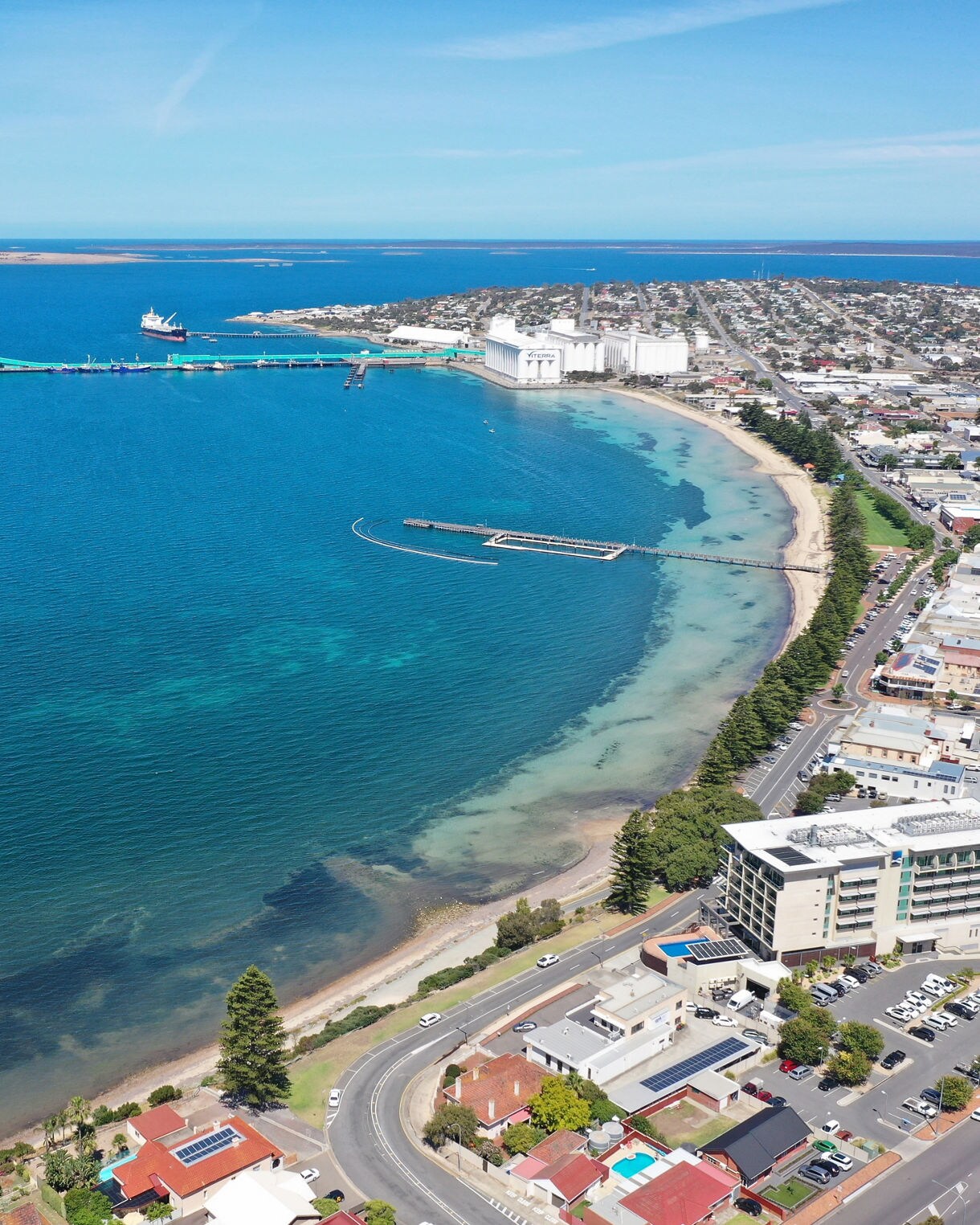 Aerial view of Port Lincoln showing a curved shoreline with clear blue water, a long pier, waterfront buildings and residential streets stretching inland.