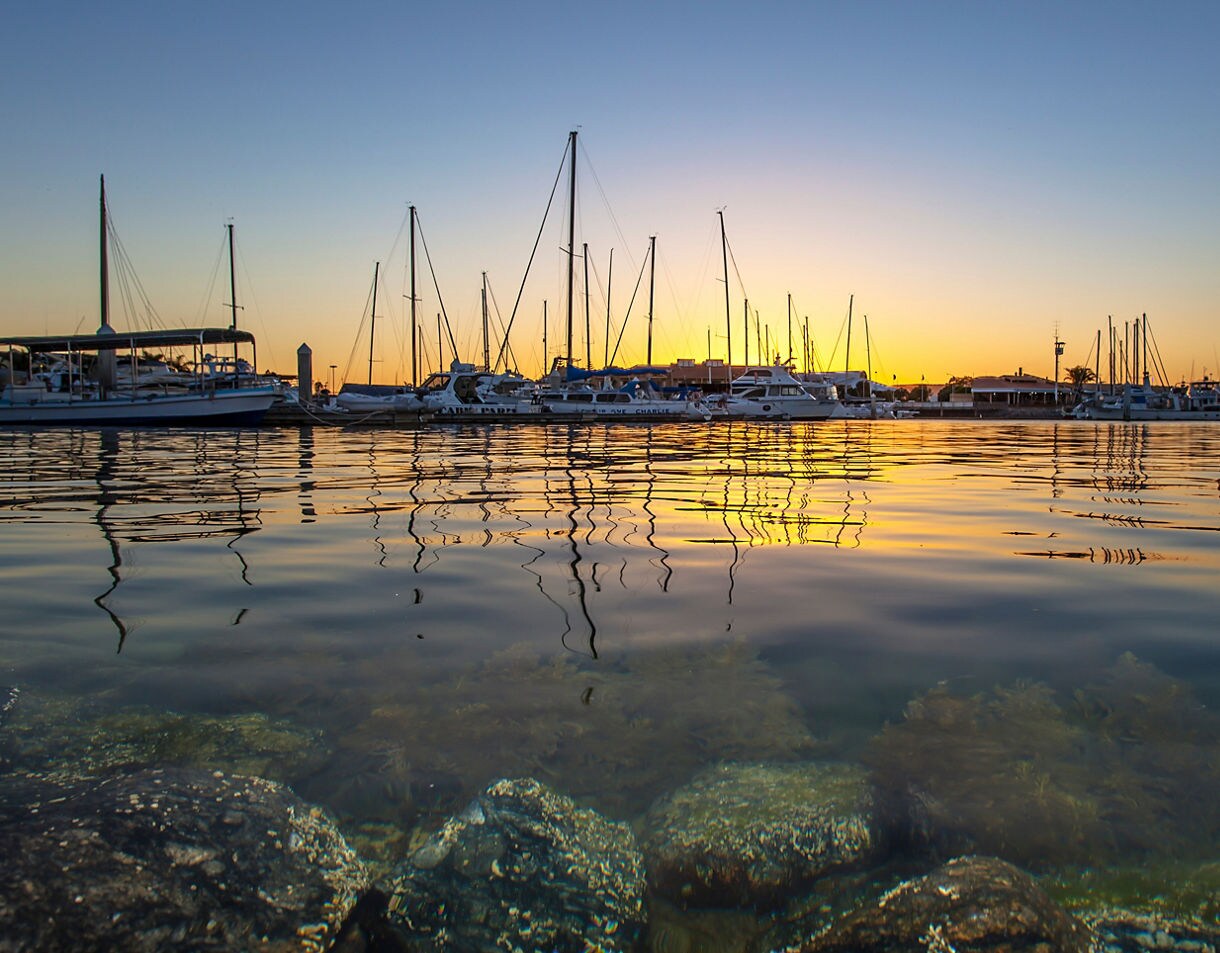 Sunset over a marina with docked sailboats reflecting on smooth golden water, with rocky shallows visible in the foreground.