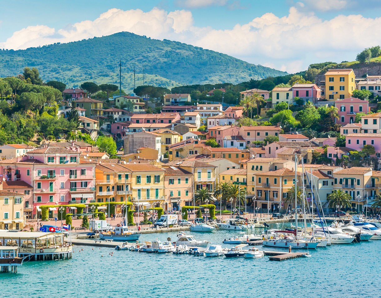 Colorful waterfront buildings in Porto Azzurro on Elba Island, with pastel houses rising up a green hillside behind a busy marina filled with boats.
