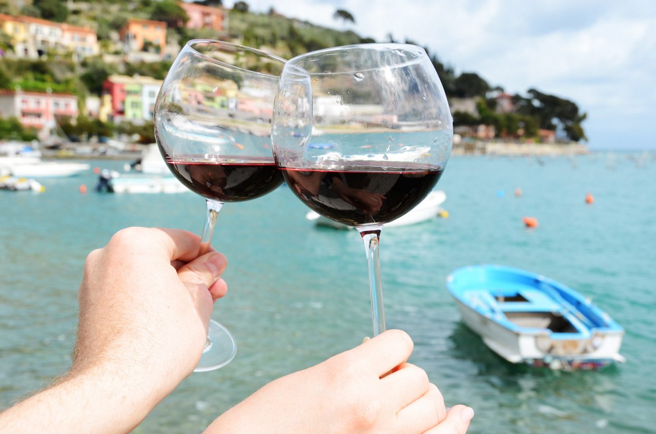 Two people clink wine glasses by the harbor in Portovenere, Italy, with colorful buildings and boats in the background.