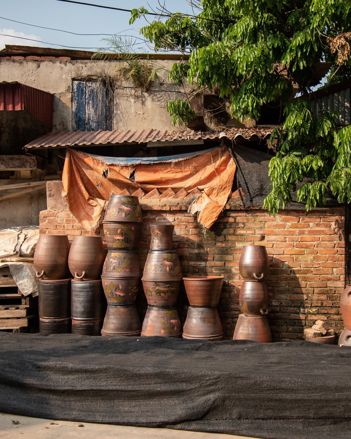 Rows of traditional clay pots stacked against a brick wall in a Vietnamese pottery village, shaded by green trees and warm sunlight.