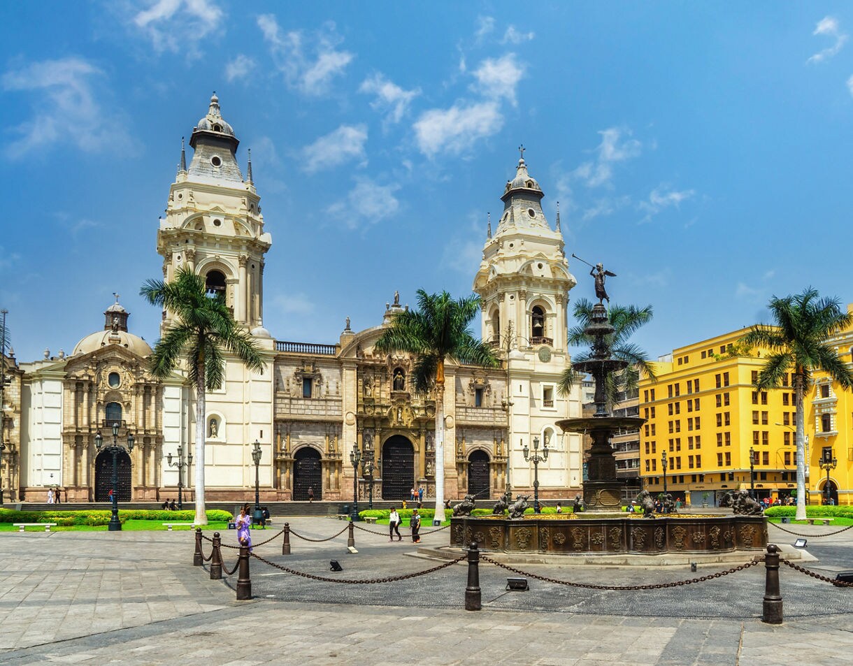 A bright city plaza with a central ornate fountain, tall palm trees and Lima Cathedral rising beside yellow colonial buildings under a clear blue sky.