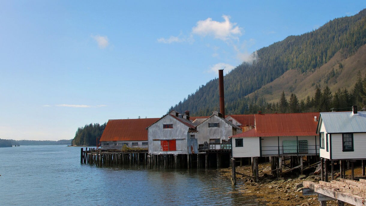 Historic cannery buildings with rusted red roofs and wooden stilts lining the waterfront at the North Pacific Cannery Historic Site.