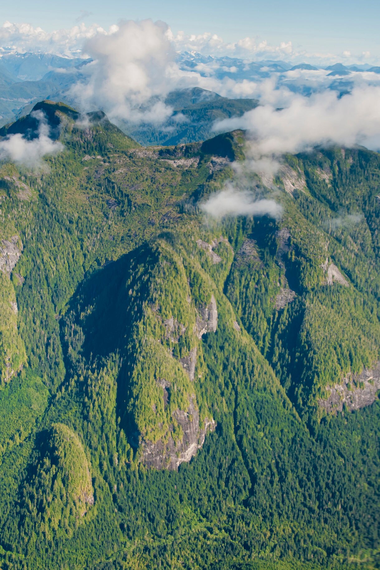 Aerial view of the Great Bear Rainforest near Prince Rupert, showing steep, forested mountains with rocky cliffs and scattered clouds drifting over the peaks.