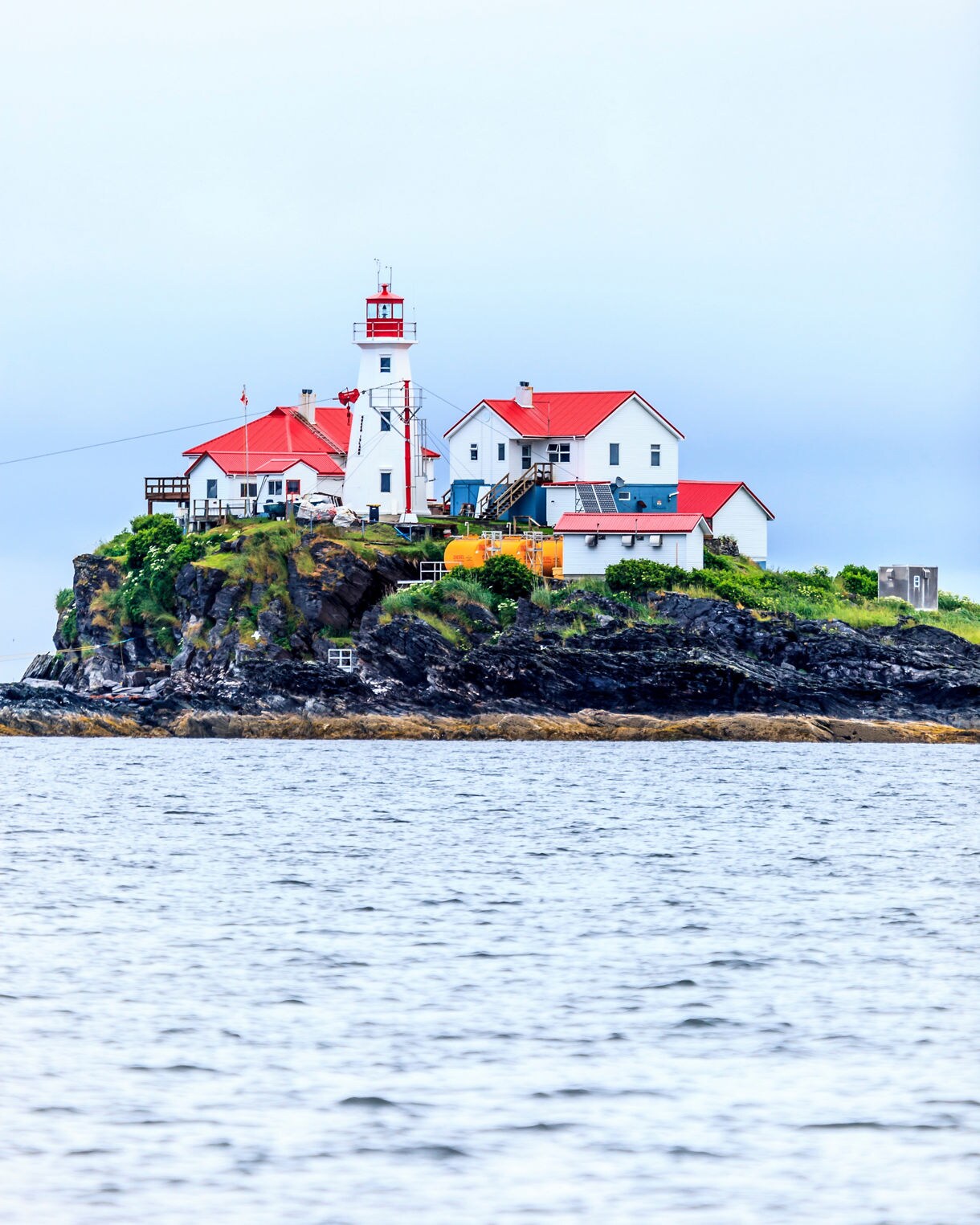 Green Island Lighthouse near Prince Rupert, British Columbia, featuring white buildings with red roofs perched on a rocky island surrounded by calm ocean water.