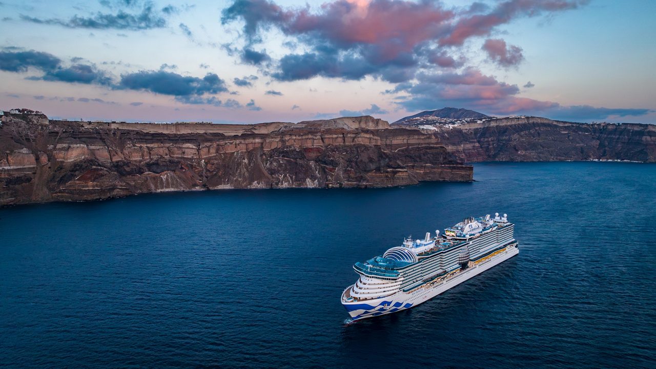 A large, modern cruise ship sails through calm blue waters near a dramatic rocky coastline at sunset. The sky features soft pink and blue hues, adding to the serene atmosphere. The ship's design is sleek and spacious, ideal for leisure travel and exploration.