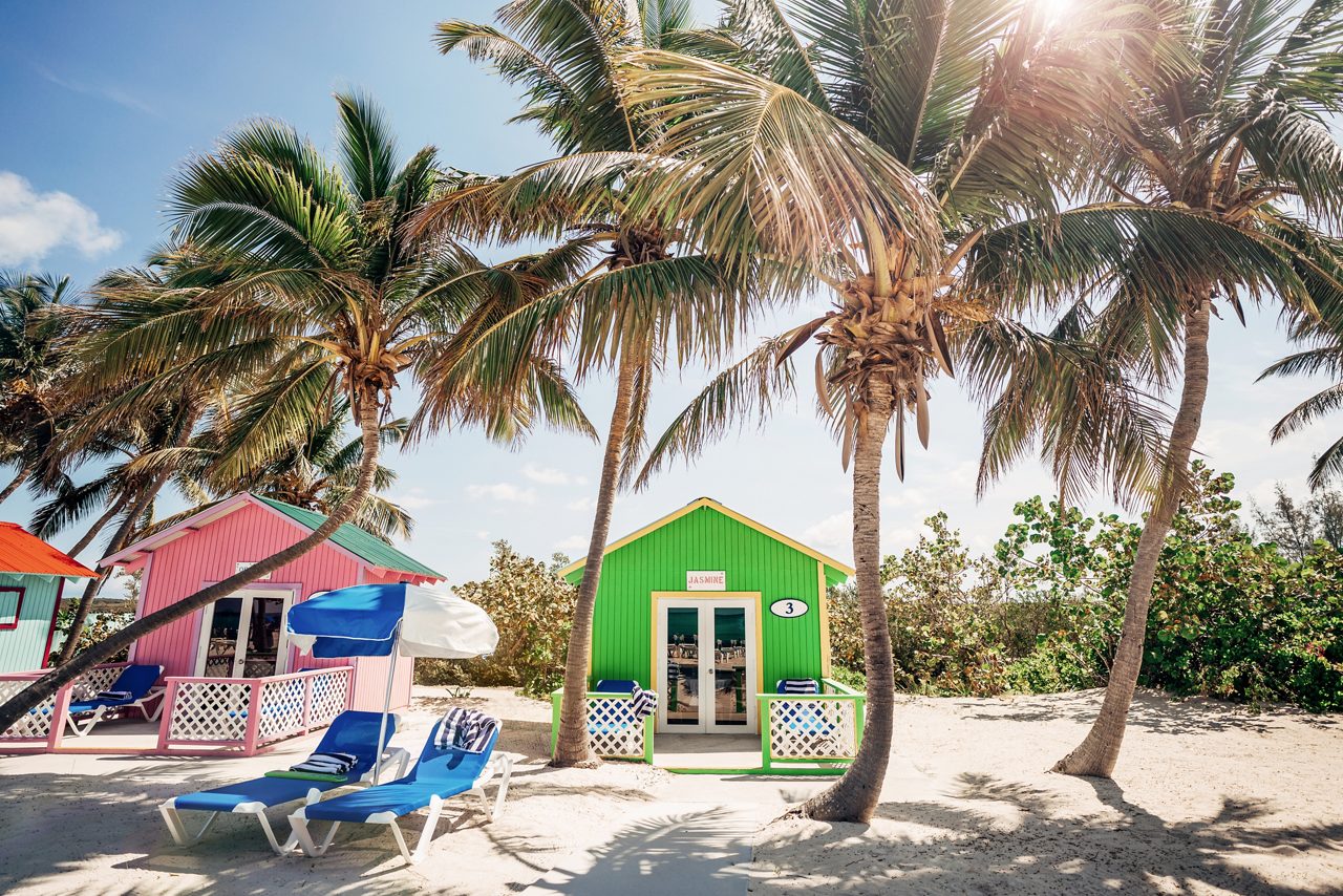 A row of vibrant, colorful cabanas on the sandy beach of Princess Cays, surrounded by palm trees and lounge chairs under bright umbrellas. 