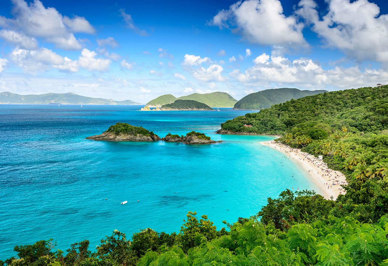 Aerial view of a tropical beach with turquoise waters, surrounded by green hills and islands under a blue sky with white clouds.