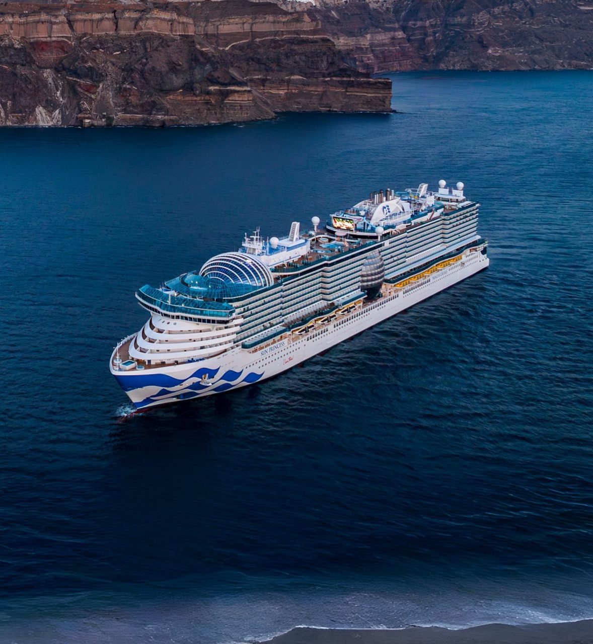 A large Princess cruise ship sails through a serene glacier bay surrounded by snow-capped mountains and lush green hills. The calm blue water reflects the stunning natural landscape under a clear sky. The image captures the grandeur of the ship and the pristine beauty of the environment.