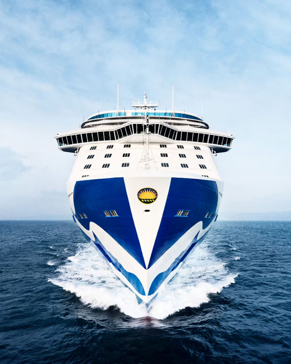 Front view of a Princess Cruises ship sailing on the ocean, with a distinctive blue and white hull design against a backdrop of clear sky and calm sea.